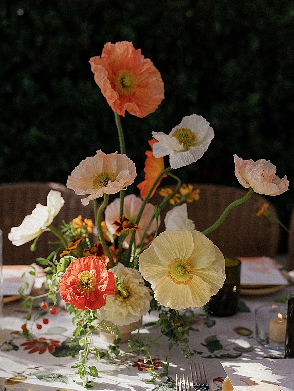Wedding centerpiece with poppy wedding centerpiece blooms in a vase, taper and votive candles, menu and place cards on floral tablecloth, dark foliage backdrop