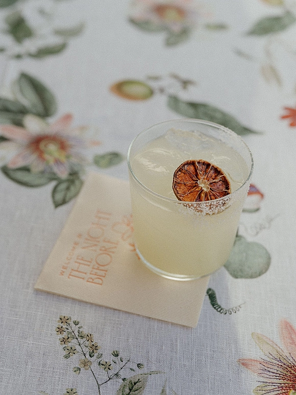 Signature wedding cocktail in a rocks glass with sugar rim and dried citrus garnish, set on a floral tablecloth with napkin