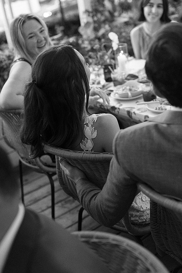 Wedding reception guests in candid conversation around a candlelit table with glassware and plates on an outdoor patio amid greenery
