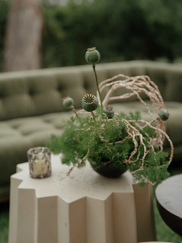 Wedding centerpiece with green wedding centerpiece foliage, poppy pods and twig branches in a black bowl on a pedestal beside a votive candle
