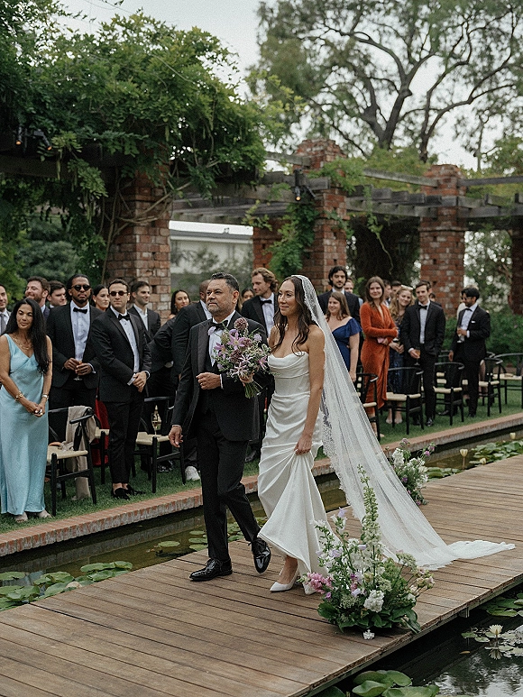Wedding processional as bride walking down aisle in strapless gown and veil, holding bouquet, with guests by a lily pond walkway and pergola backdrop