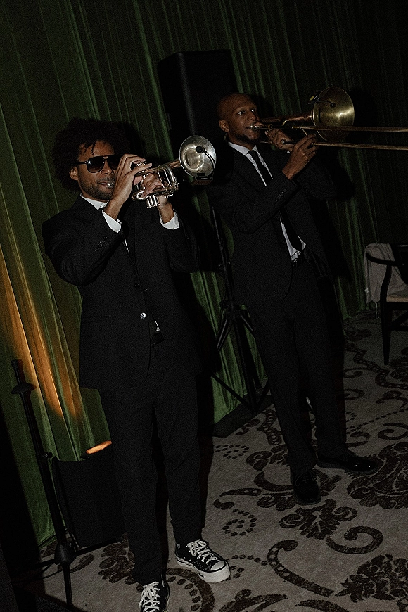 Wedding band brass duo in black suits and sunglasses playing trumpet and trombone by green drapery with microphone stand indoors