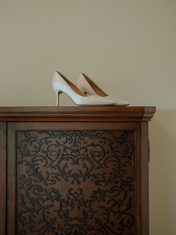 Bridal shoes, white wedding heels perched on a wood dresser against a neutral wall with carved cabinet detailing