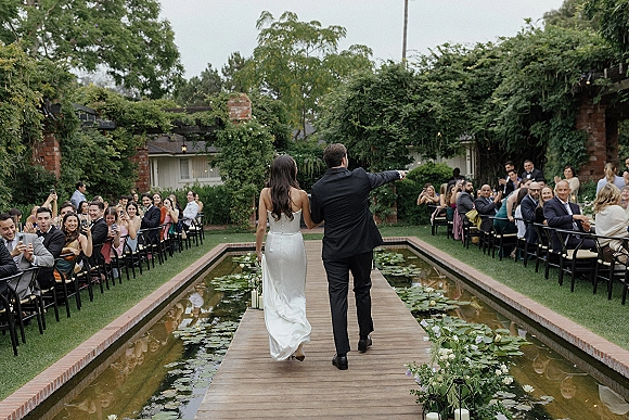 Wedding recessional as bride and groom walk a wood bridge aisle over a lily pond, guests cheering amid lanterns, candles, and string lights