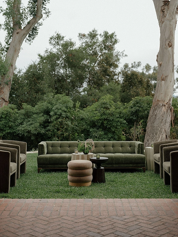 Lounge seating area with velvet sofa and upholstered armchairs around candles and bud vase florals on a brick patio lawn under trees