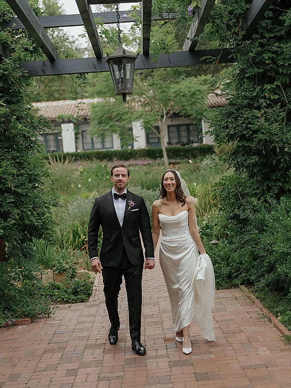 Couple portrait of bride and groom walking hand in hand under a pergola with a hanging lantern, lush garden greenery and brick walkway behind