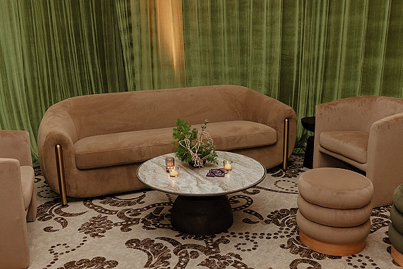 Wedding lounge area with a velvet sofa and upholstered armchairs around a round marble coffee table, candles, and greenery before green drapes