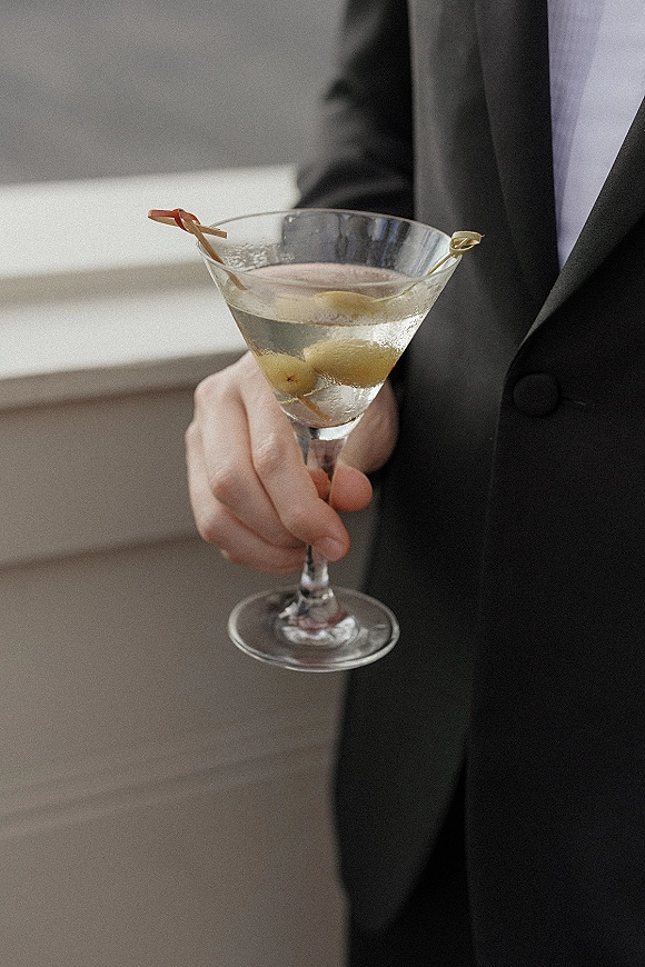 Martini cocktail in a martini glass with olives on a pick, held by a suited hand in tie under soft window light indoors