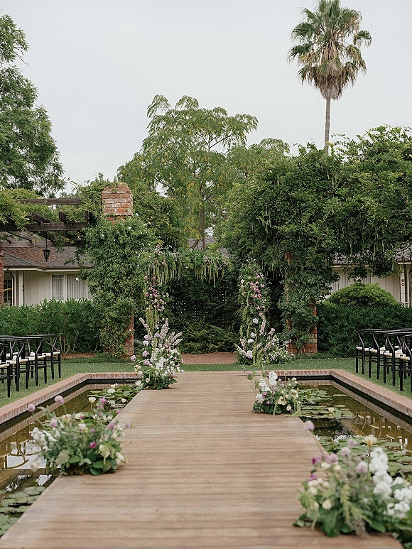 Ceremony aisle design with garden ceremony aisle flowers lining a wooden walkway over a lily pond, leading to a floral arch by brick columns