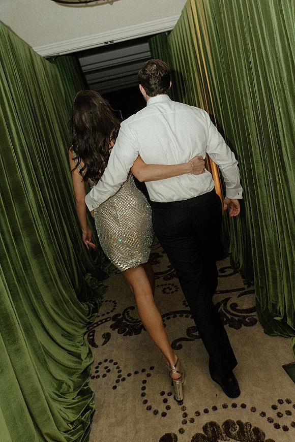 Couple exit as newlyweds walk away arm in arm, bride in a sequined mini dress and heels, groom in white shirt by green velvet drapes