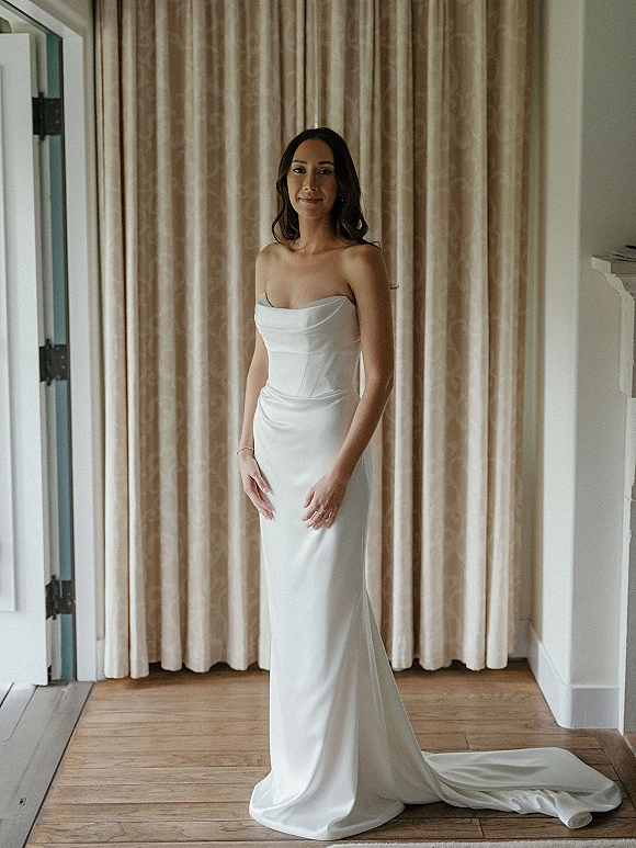 Bridal portrait of a bride in a strapless wedding dress with draped neckline, bracelet and ring, standing by beige curtains indoors