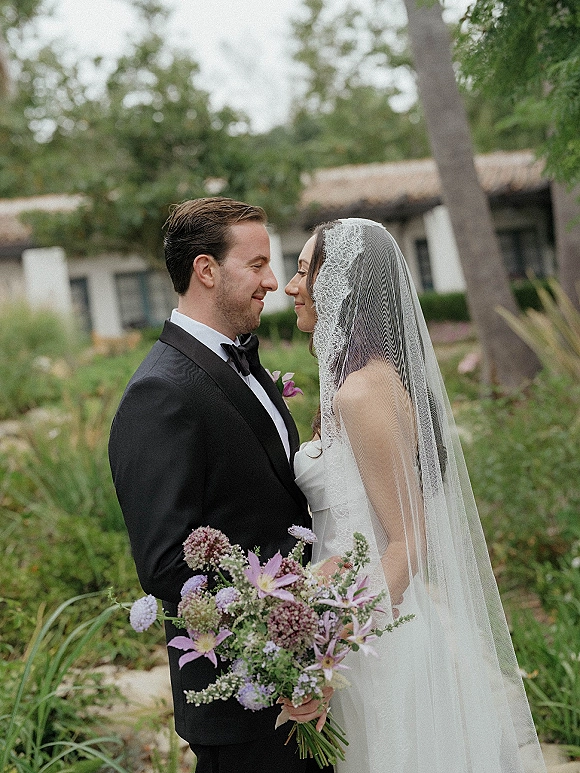 Couple portrait of bride and groom nose to nose, her lace veil and purple bouquet against garden greenery by a stone path