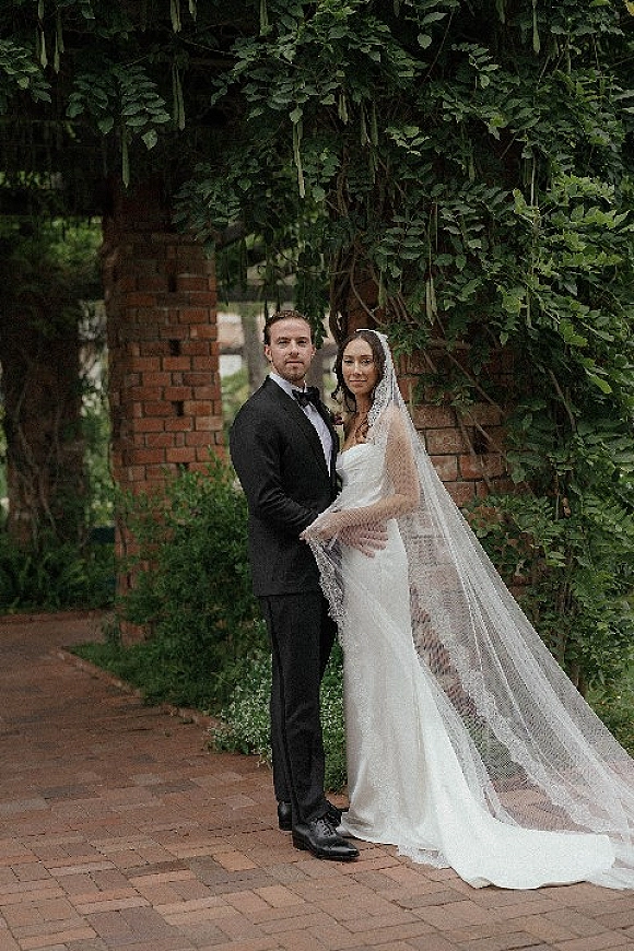 Couple portrait of bride and groom pose under a vine-covered arbor, bride in a strapless dress with veil beside groom in tuxedo on brick walkway