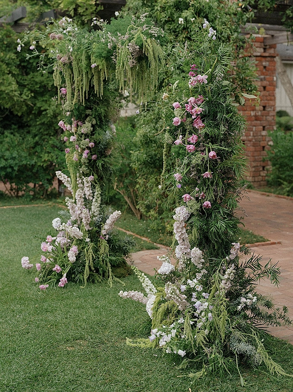 Wedding ceremony arch with hanging amaranthus, pink and white blooms, and greenery garland on a garden lawn beside a brick walkway