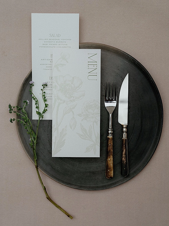 Wedding tablescape with wedding place setting featuring a menu card, dark charger plate, wood-handled flatware, and floral sprig on neutral linen