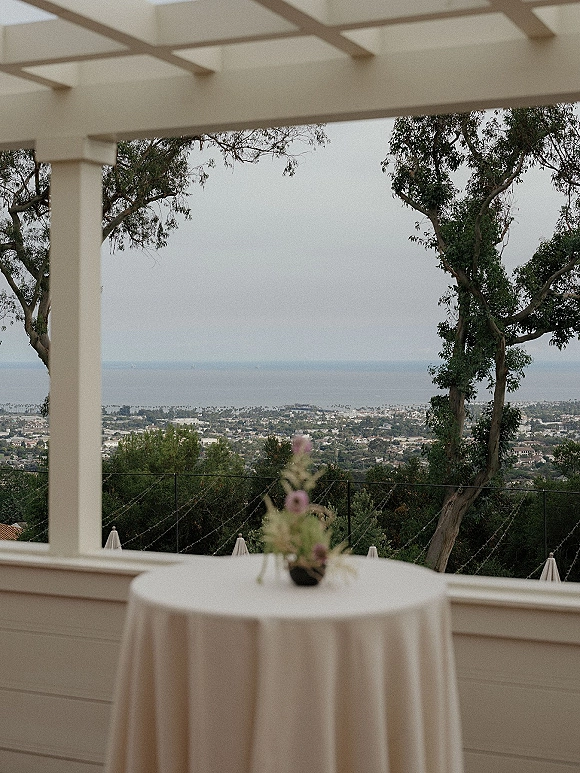 Cocktail table decor with a wedding cocktail hour table draped in ivory linen and a bud vase centerpiece on a pergola terrace with ocean view