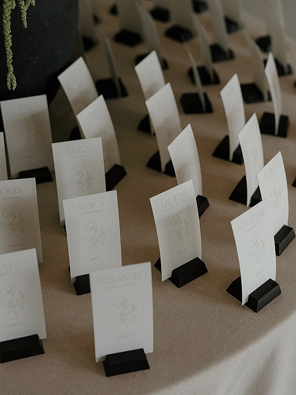 Wedding escort cards in white tent cards with botanical line art, set in black holders with guest names and table numbers on linen tabletop