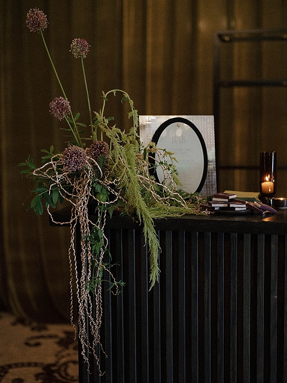 Wedding welcome table with acrylic welcome sign and asymmetrical florals, pillar candles and greenery against a wood-paneled wall