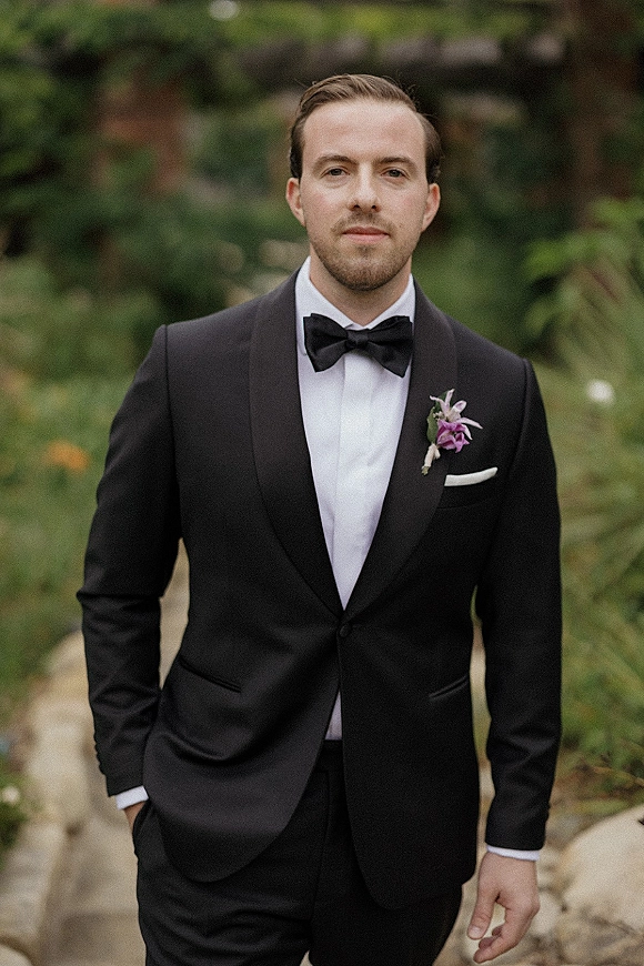Groom portrait in a black tuxedo groom look with bow tie and boutonniere, hands in pockets on a stone path amid garden greenery
