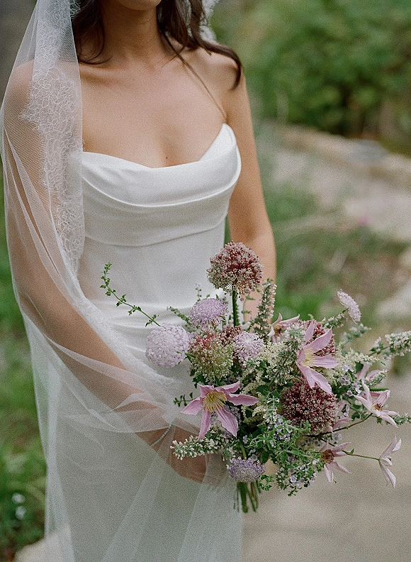 Bridal portrait of a bride holding bouquet with a strapless satin wedding dress and lace-edge veil on a lush garden path