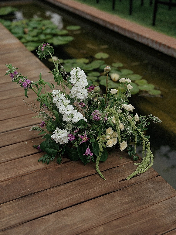 Wedding bouquet with garden style blooms of white and purple flowers, cream roses and greenery on a wooden dock by lily pads on pond