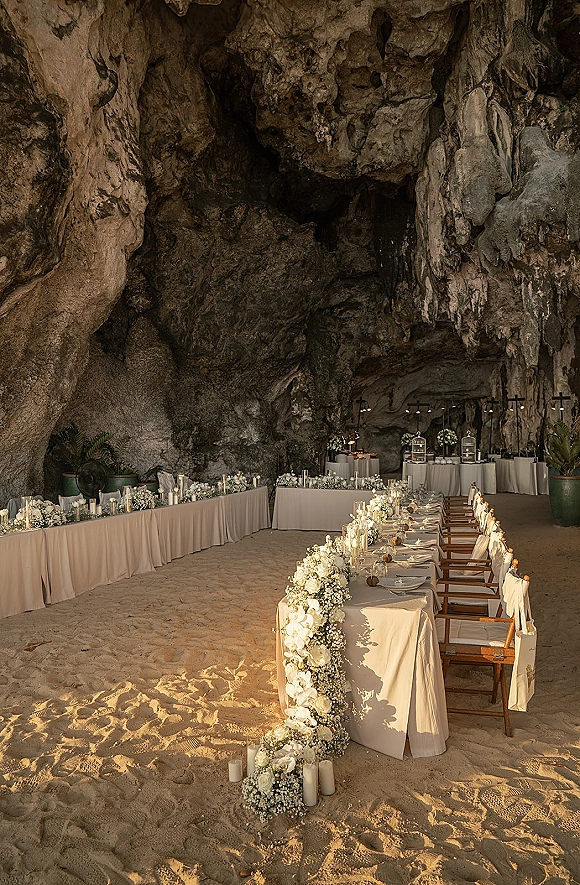 Reception tablescape with a long banquet table, white floral garland and pillar candles set inside a cave with rock walls and stalactites
