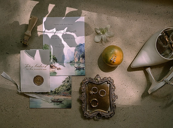 Wedding invitation suite with wax seal and ribbon, styled with rings, heels, perfume and orange on a stone surface in soft shadows