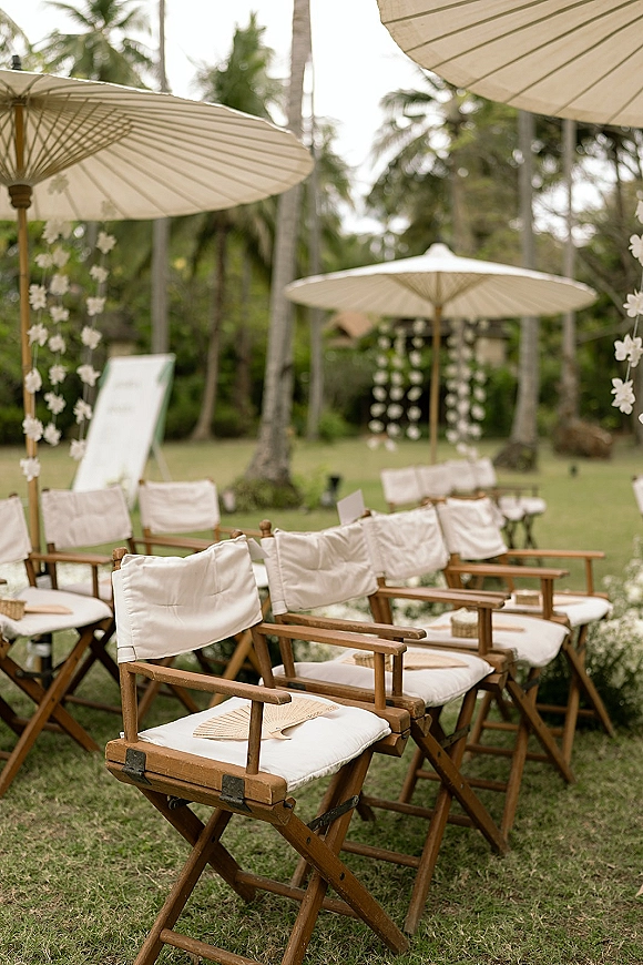 Ceremony seating with outdoor ceremony chairs, white cushions and paper parasols on a grass lawn framed by palm trees and tropical greenery
