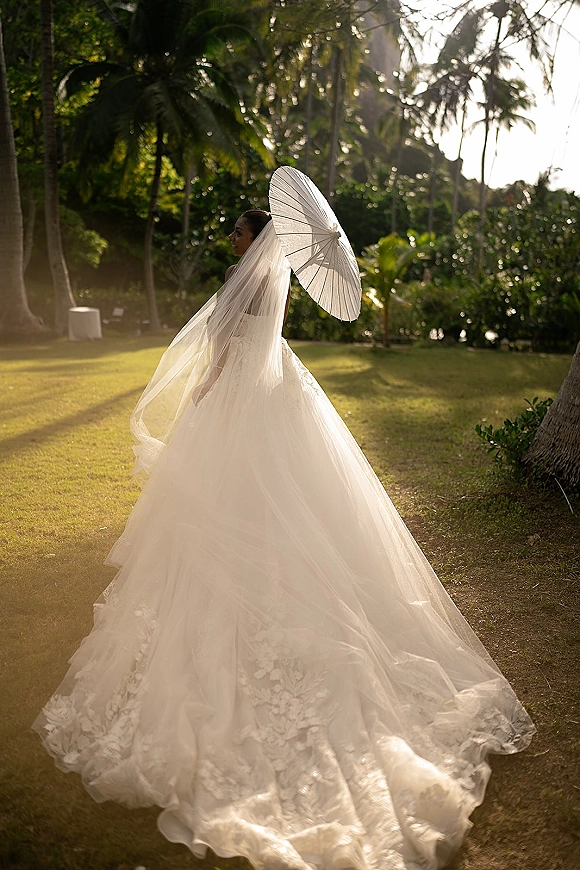 Bridal portrait of a bride with parasol in a strapless wedding gown and cathedral veil, backlit on a palm-lined tropical lawn at sunset
