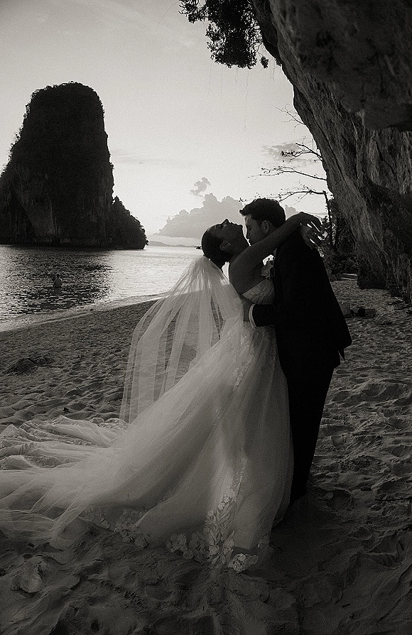 Wedding kiss portrait in black and white of bride and groom embracing on beach sand, veil flowing by ocean and rocky cliffs under cloudy sky