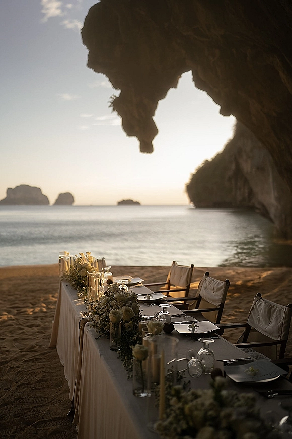 Beach reception tablescape with destination wedding reception details, white floral garland and taper candles on a long table by ocean cliffs at sunset