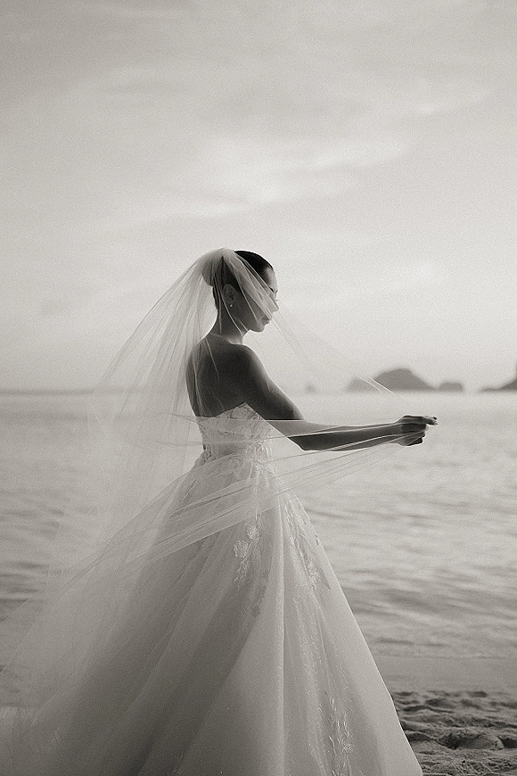 Bridal portrait of a bride holding her veil, wearing a strapless lace gown and earrings on a beach shoreline with ocean and distant islands