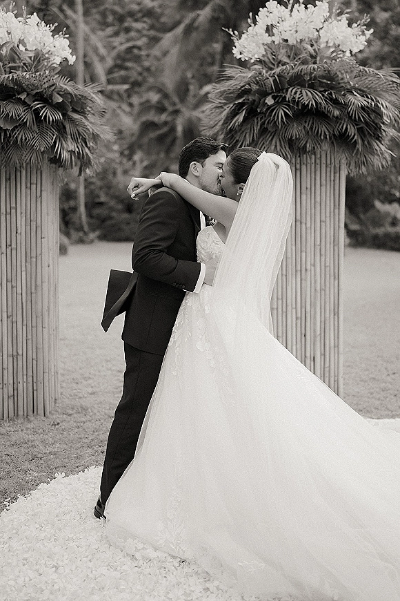 Wedding kiss portrait of the bride and groom kissing under a floral bamboo arch, her veil flowing over a strapless lace dress in a garden setting