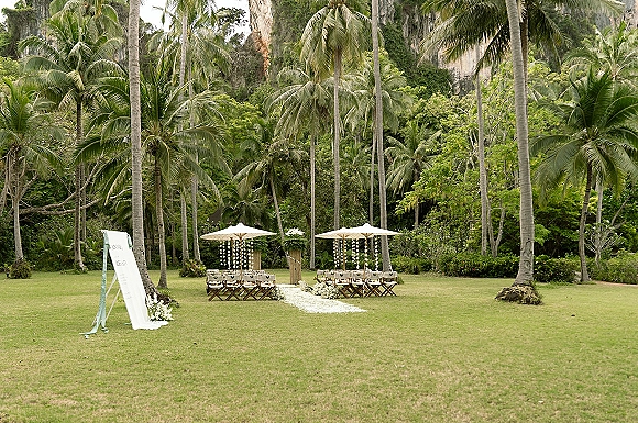 Outdoor ceremony setup with tropical wedding ceremony seating, white aisle runner and hanging florals beneath palm trees on a cliffside lawn