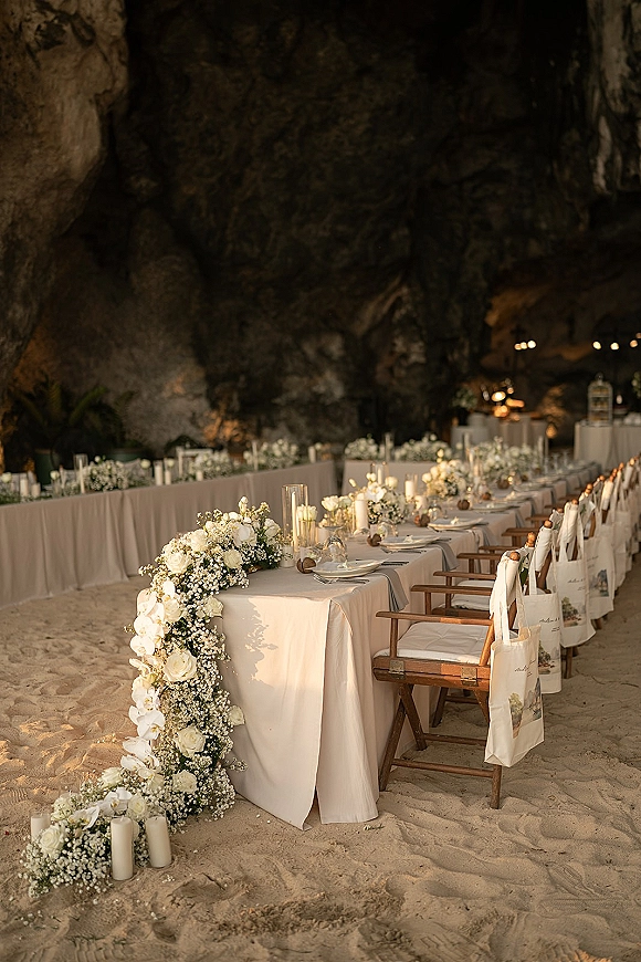 Reception tablescape on a long banquet table wedding with white floral garland and candlelight, set on sand inside a cave with rock walls