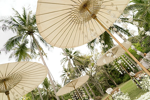 Ceremony decor with outdoor wedding ceremony aisle lined with white florals, wooden benches, and parasols beneath palm trees on a lawn