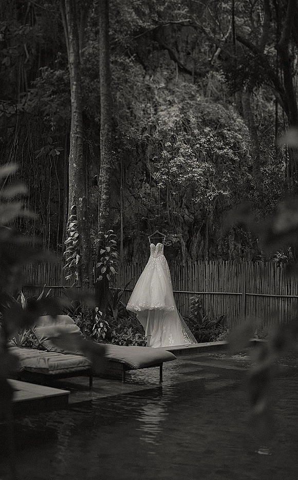 Wedding dress, lace wedding dress hanging on a hanger with long veil beside a pool, framed by tropical greenery and a bamboo fence
