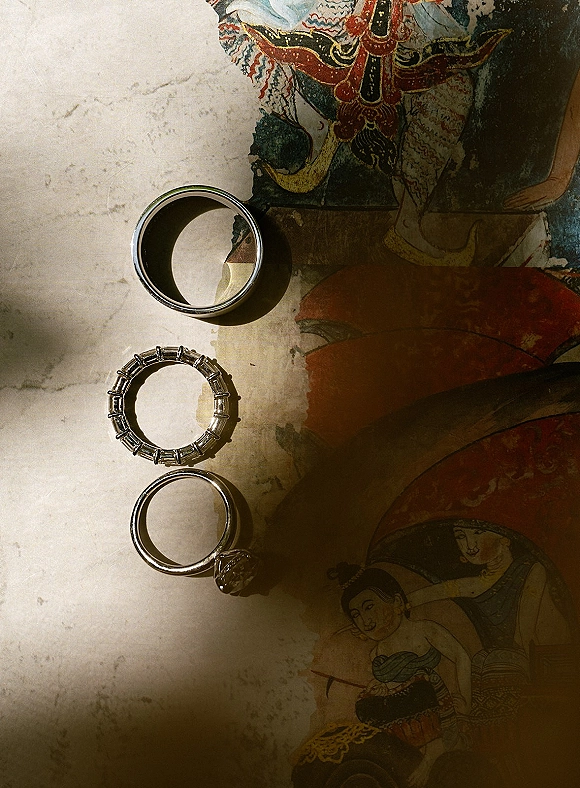 Wedding rings flat lay featuring an engagement ring and band stack with a diamond accent on patterned artwork, casting moody shadows