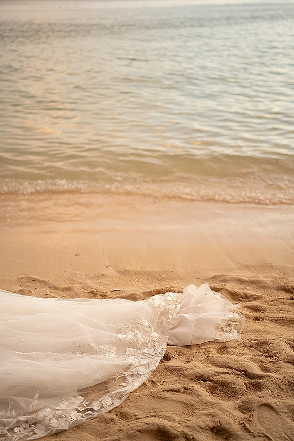 Wedding dress train with lace applique trailing across sandy beach near the shoreline, tulle fabric catching light beside ocean water
