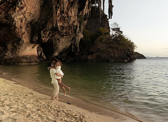 Couple portrait on a beach engagement photo, hugging by ocean water near a rocky cliff and sea cave, her white dress and his linen outfit