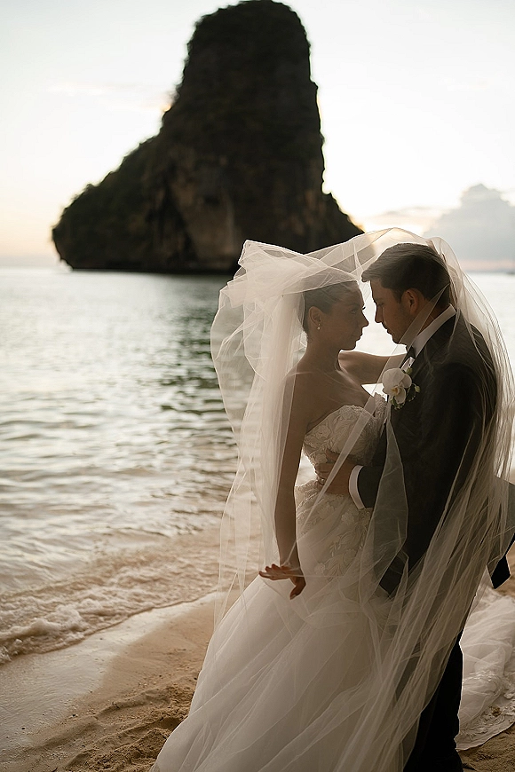 Couple portrait at a beach wedding with a veil draped over them, bride in lace dress and groom in tuxedo by ocean rocks