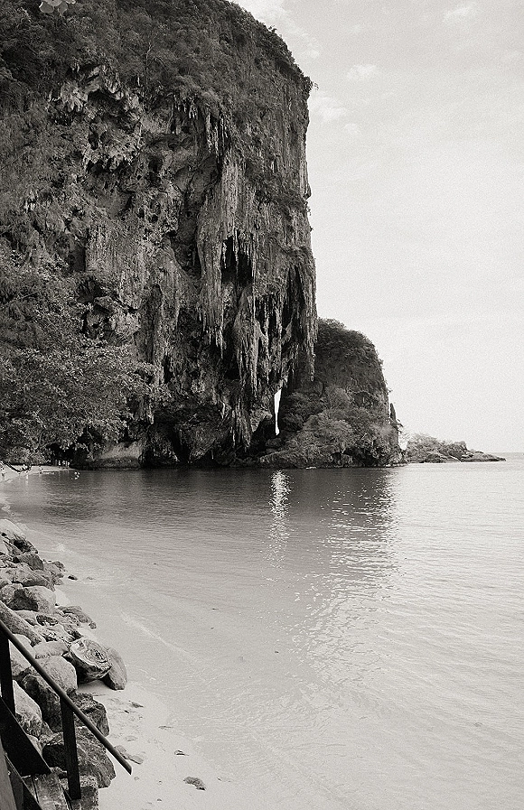 Beach cliff landscape in a black and white seascape, showing a rocky cliff and shoreline rocks beside calm ocean water and sandy beach under open sky