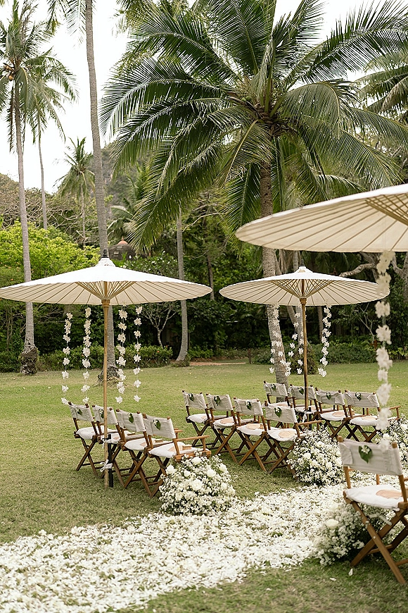 Ceremony setup for an outdoor wedding ceremony with white parasols, petal aisle, floral clusters and wooden chairs on a tropical lawn with palms