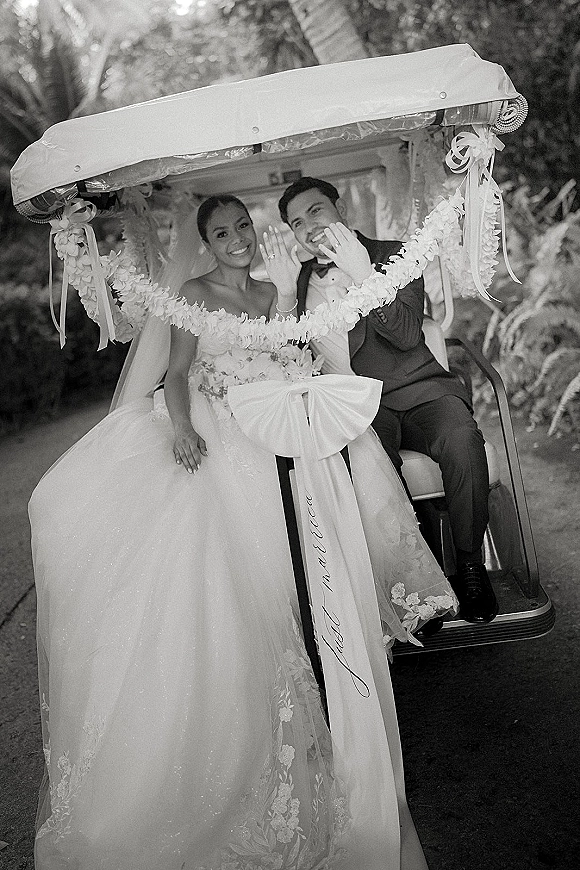 Couple portrait of newlyweds in a just married golf cart, smiling and showing wedding rings along a palm-lined path in tropical greenery