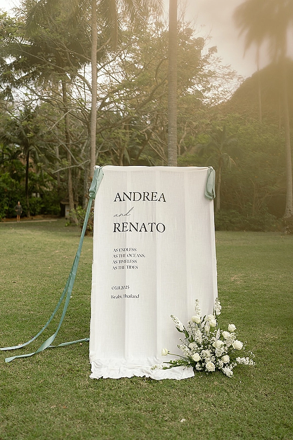 Wedding welcome sign with serif typography on fabric, ribbons, and white roses with baby’s breath on a tropical garden lawn with palms