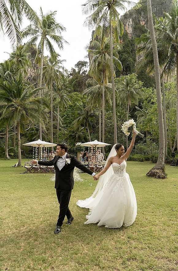 Wedding recessional as bride and groom walk away holding hands, bride raising bouquet along petal-strewn aisle with palms and umbrellas