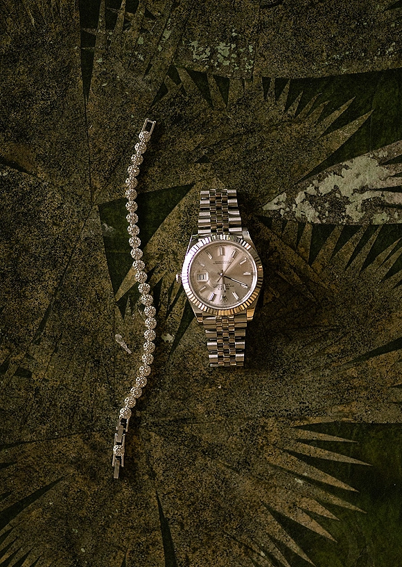 Groom accessories featuring a wedding day watch beside a diamond tennis bracelet on a patterned green and gold surface flat lay