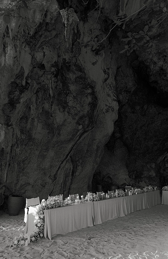 Reception head table with a wedding sweetheart table floral garland and candlelight on white linen, set against a rock cave wall and sand floor