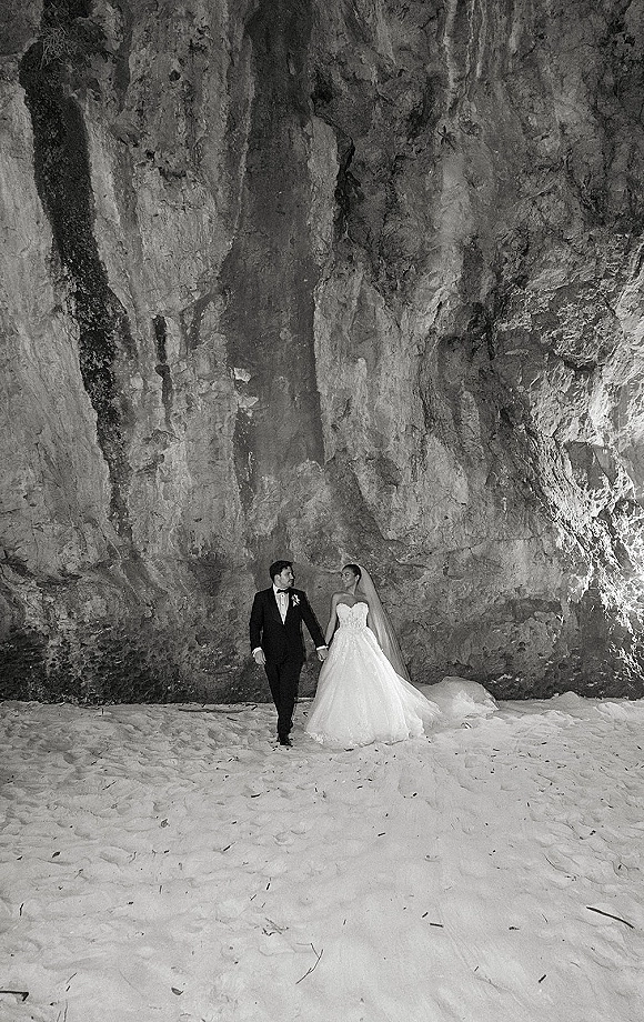 Couple portrait in a black and white wedding portrait, bride in ball gown and long veil holding groom’s hand on sand beach by rock cliff