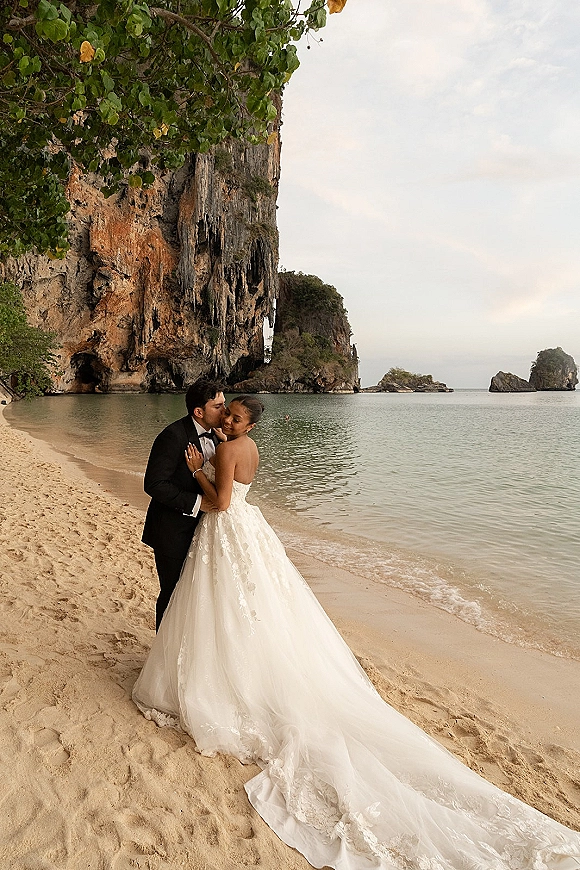Wedding couple portrait on a sandy beach, groom kissing bride’s forehead as her strapless lace dress train flows by ocean cliffs